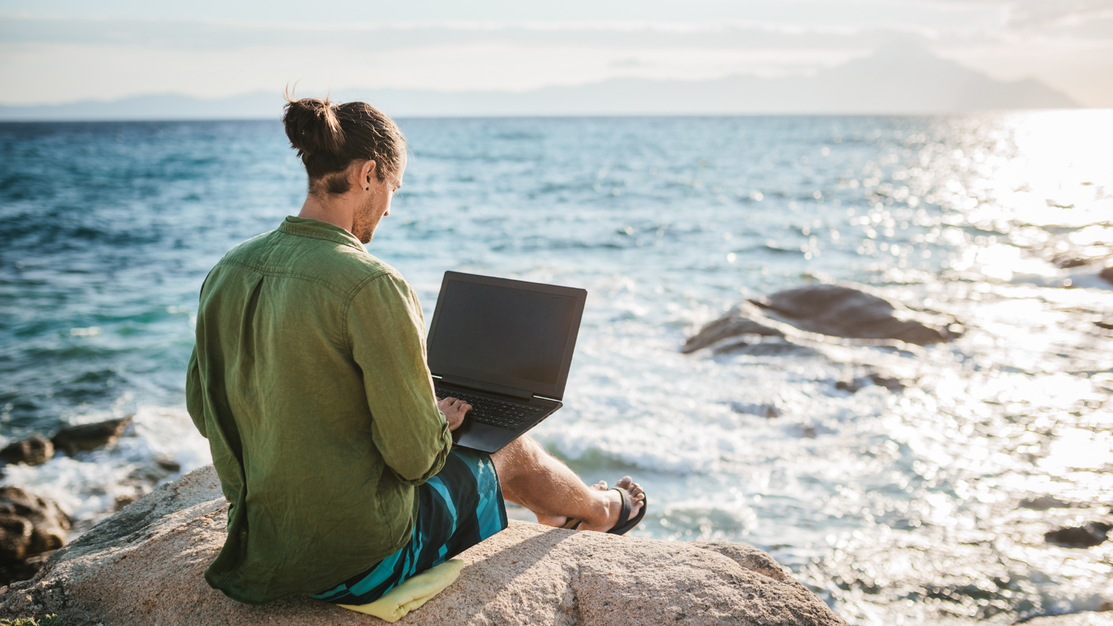 Eco nomad working on a laptop by the ocean at sunset, combining remote work with sustainable travel lifestyle