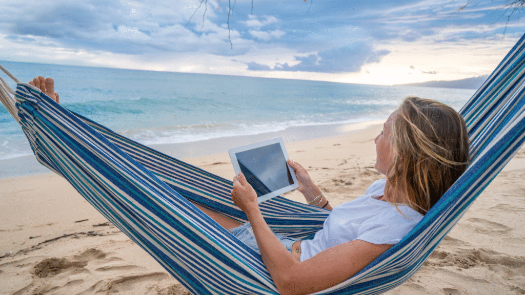 Woman working on a laptop from a hammock on a quiet tropical beach under palm shade.
