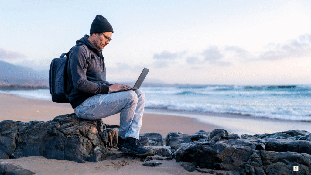 Woman working on a laptop on a rocky beach, practicing green travel as a digital nomad in 2025