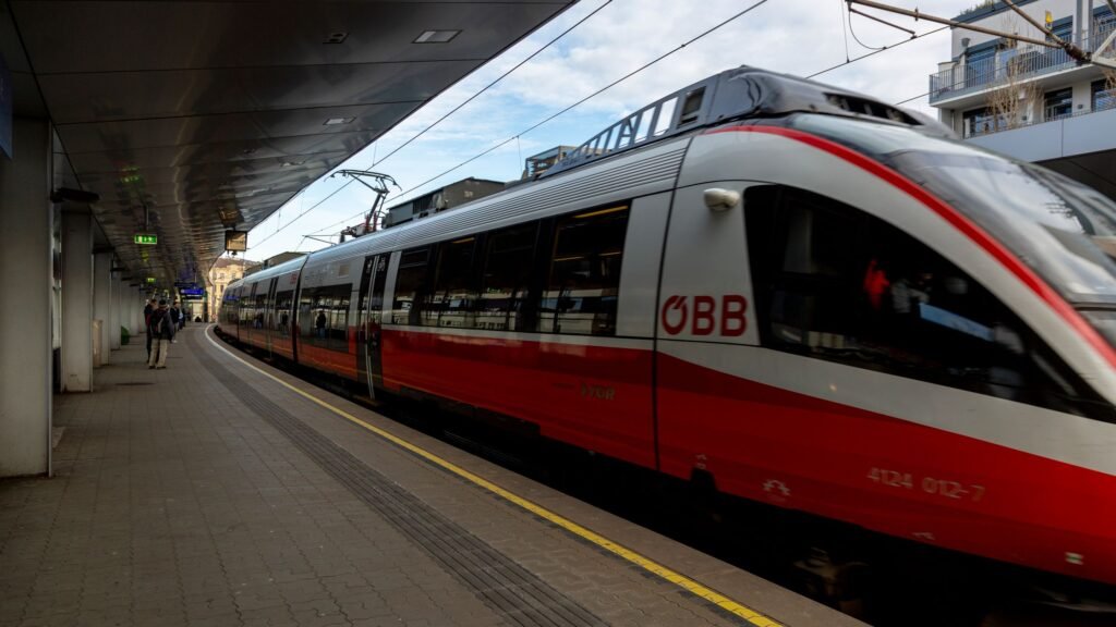 A sleek red and white ÖBB train on the platform at a modern station, with passengers waiting.